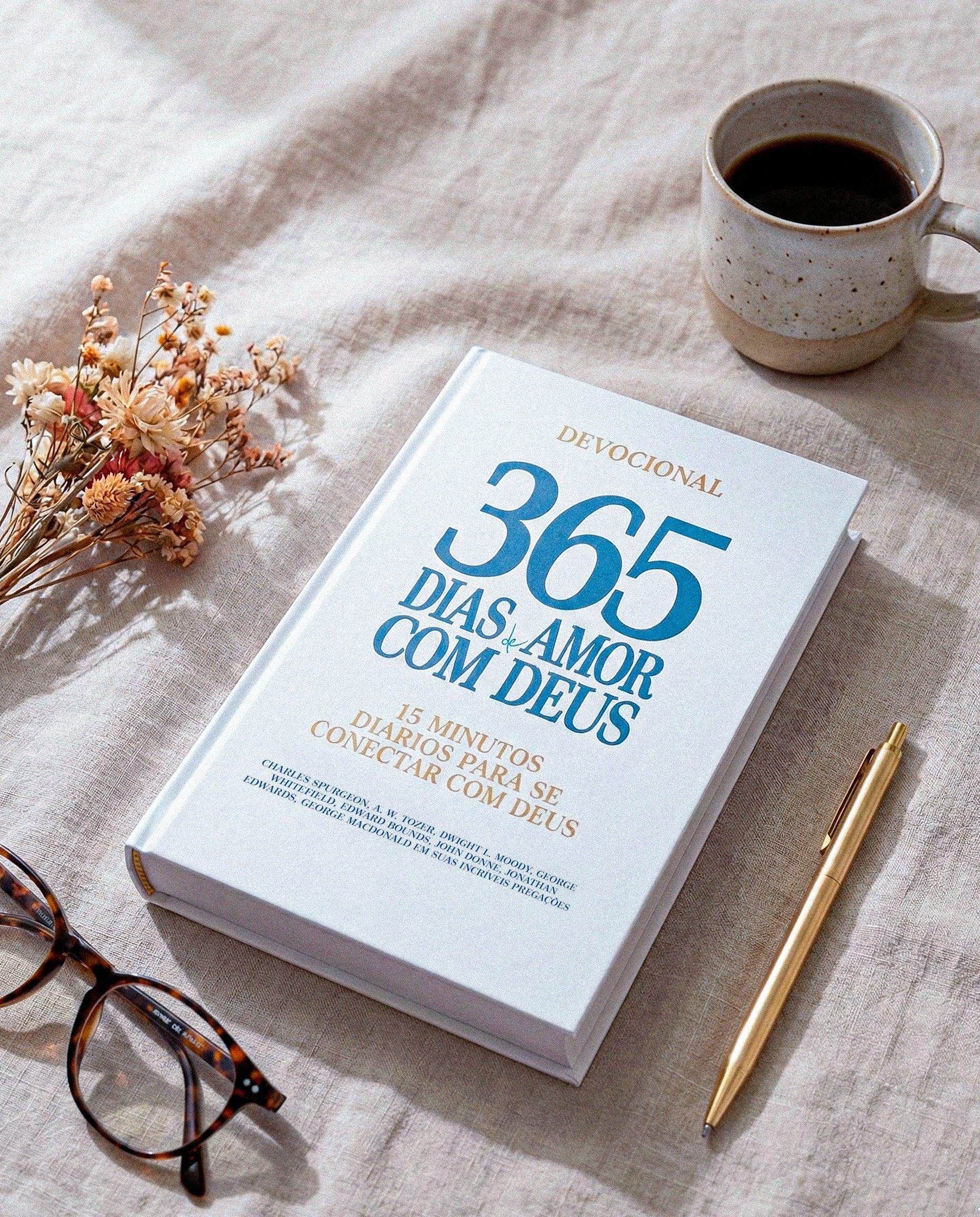 Livro devocional 365 dias de amor com Deus, caneta dourada, óculos e xícara de café sobre tecido claro